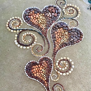 Op de afbeelding: Een hartvormig stenen kunstwerk met drie harten en wervelende accenten. De harten zijn gevuld met kleine, veelkleurige stenen en de wervelingen zijn gemaakt van witte en bruine stenen. Het kunstwerk staat op een zandkleurig oppervlak, met een steen "Beach Art".