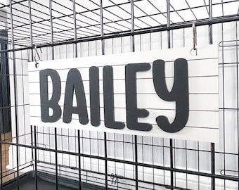 Könnte beinhalten: Ein weißes Holzschild mit dem Namen "BAILEY" in fetten, schwarzen Buchstaben. Das Schild ist an einer schwarzen Draht-Hundebox befestigt. Der Hintergrund ist eine hellfarbige Wand.