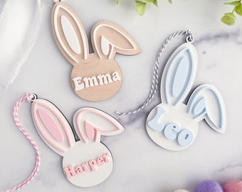 May include: Three wooden bunny-shaped ornaments with personalized names. The bunny ornaments are in pink, blue, and brown with white accents. The names are "Emma", "Leo", and "Harper".