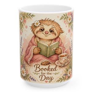 May include: White ceramic mug featuring a watercolor illustration of a sloth reading a book while wrapped in a pink blanket. The mug has the text "Booked for the Day" and is decorated with flowers and a teacup.