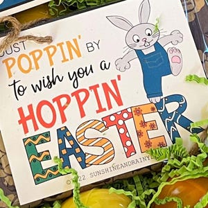 Può includere: Un biglietto di Pasqua colorato con un coniglio che salta sulla parola "Pasqua". Il biglietto dice "Just poppin' by to wish you a hoppin' Easter".