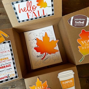 May include: A brown cardboard box filled with autumn-themed items, including a colouring page with a large orange leaf, a pumpkin spice latte cup, and several autumn-themed cards with text like "hello fall", "football games", and "you are un-de-leafable".