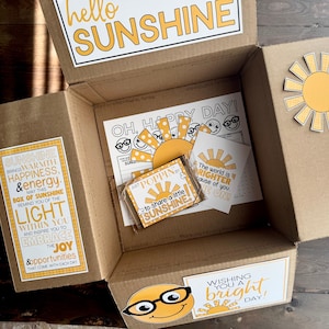Può includere: Una scatola di cartone piena di oggetti a tema sole. La scatola contiene biglietti con grafiche solari e frasi come "Hello Sunshine" e "Wishing you a bright day!" La scatola ha anche un adesivo a forma di sole.