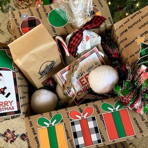 Könnte beinhalten: Ein brauner Karton, gefüllt mit Weihnachtsgeschenken. Die Schachtel ist mit rot-schwarzem kariertem Papier, grün-rotem Band und verschiedenen Weihnachtsornamenten verziert. Zu den Geschenken gehören eine Tüte Kaffee, ein Zuckerstange, ein Schneemann-Anhänger und ein Geschenkanhänger mit der Aufschrift "Frohe Weihnachten".