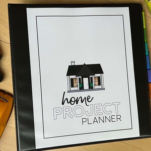 Puede incluir: Una carpeta negra con una cubierta blanca que dice "home PROJECT PLANNER" en texto negro. La cubierta también tiene una ilustración en blanco y negro de una casa. La carpeta tiene pestañas de colores en el lateral.
