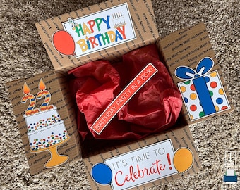 May include: A brown cardboard box with colorful birthday decorations, including a cake with candles, balloons, and the text "Happy Birthday" and "Birthday Party in a Box!" The box is filled with red tissue paper.