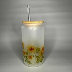 May include: A frosted glass tumbler with a bamboo lid and a clear glass straw. The tumbler is decorated with yellow sunflowers and green leaves. The tumbler is approximately 6 inches tall.