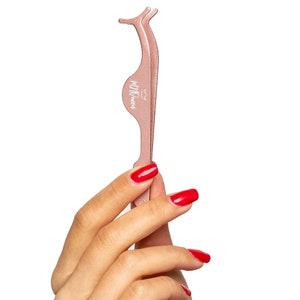 Op de afbeelding: Wimperapplicator-gereedschap van rosé goud met de woorden "lil' queen" erop gedrukt. Het gereedschap wordt vastgehouden door een hand met rode nagellak.