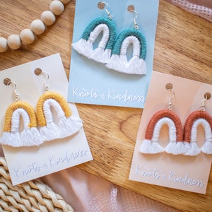 May include: Three pairs of rainbow-colored crocheted earrings with white fringe on a wooden surface. Each pair is displayed on a card with the text "Knots of Kindness".