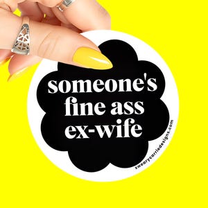 May include: A round white sticker with black text that reads "someone's fine ass ex-wife" in a black cloud shape. The sticker is held by a hand with yellow nails and silver rings, set against a bright yellow background.