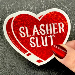 Pu&ograve; includere: Un adesivo a forma di cuore con sfondo glitterato rosso e bordo bianco. L'adesivo presenta le parole "SLASHER SLUT" in grassetto bianco. Il sito web "wearycarriedesigns.com" &egrave; stampato sul lato sinistro.