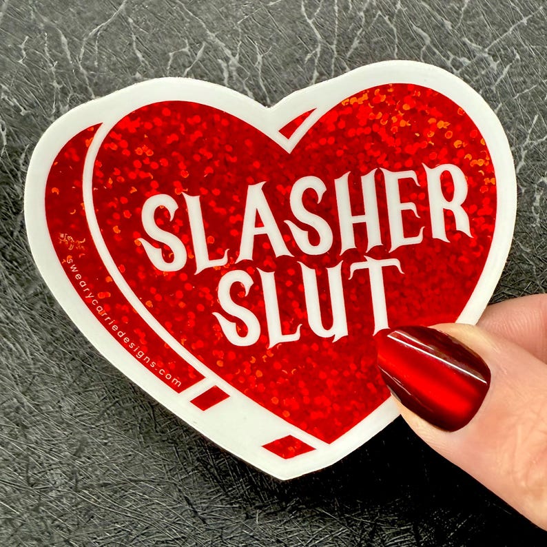 Pu&ograve; includere: Un adesivo a forma di cuore con sfondo glitterato rosso e bordo bianco. L'adesivo presenta le parole "SLASHER SLUT" in grassetto bianco. Il sito web "wearycarriedesigns.com" &egrave; stampato sul lato sinistro.