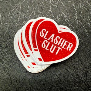 Pu&ograve; includere: Una pila di adesivi a forma di cuore con sfondo rosso glitterato e la scritta bianca "SLASHER SLUT". Gli adesivi hanno un bordo bianco e sono impilati su una superficie grigio scuro strutturata.