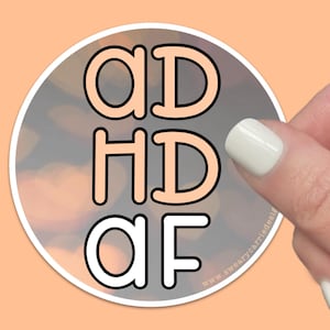 May include: A round sticker with a peach background and a white outline. The sticker says "ADHD AF" in large, bold, peach-colored letters with black outlines.