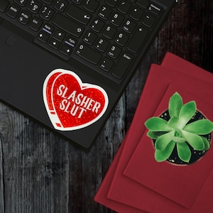 Pu&ograve; includere: Un adesivo rosso a forma di cuore con la scritta bianca "SLASHER SLUT" su un laptop nero. L'adesivo ha un aspetto usurato. Libri rossi e una pianta grassa in vaso sono accanto al laptop.
