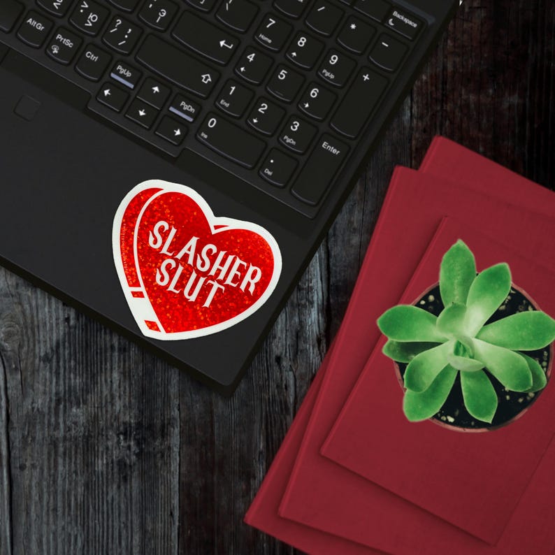 Pu&ograve; includere: Un adesivo rosso a forma di cuore con la scritta bianca "SLASHER SLUT" su un laptop nero. L'adesivo ha un aspetto usurato. Libri rossi e una pianta grassa in vaso sono accanto al laptop.