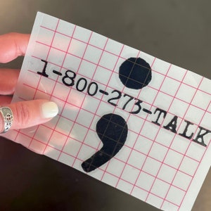 May include: A white sheet with a black semicolon and the text "1-800-273-TALK" printed on it.