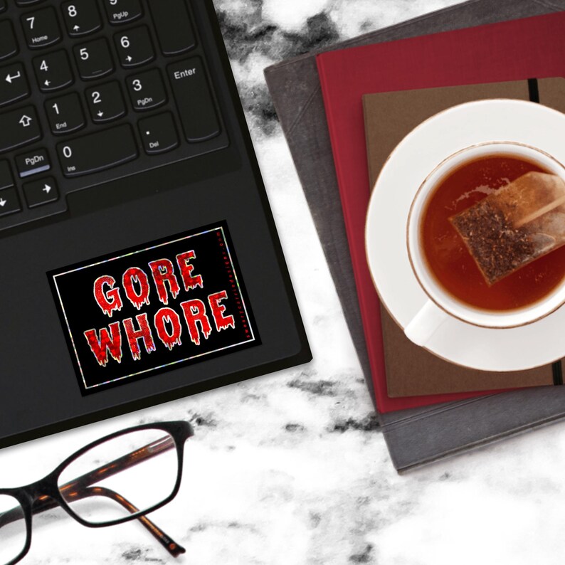 May include: A black and red sticker with the text "GORE WHORE" on a laptop computer. The sticker is on the touchpad of the laptop. There is a cup of tea and a stack of books in the background.