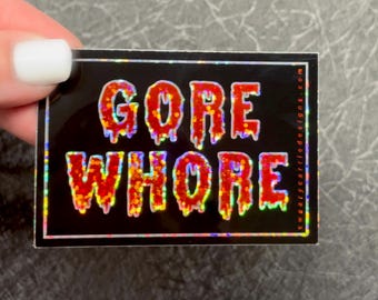 May include: A black sticker with a holographic finish features the text "GORE WHORE" in red dripping letters. The text is surrounded by a black border.