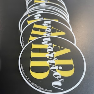 May include: A stack of round black and yellow stickers with the text "DAD" in yellow and "Sweary Carrie Designs.com" in white.