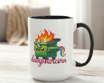May include: White ceramic mug with a black interior and handle. The mug features a cartoon illustration of a green dumpster with a unicorn horn, rainbow tail, and flames, with the word "dumpstercorn" in pink script.