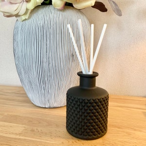 May include: A black glass diffuser bottle with a textured diamond pattern and white reeds. A large vase with black and white stripes and flowers is in the background. The objects are on a wooden surface. The bottle is for home fragrance.