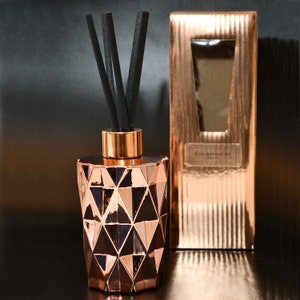 May include: A rose gold geometric diffuser bottle with black reeds. The bottle has a faceted design and a metallic gold collar. A matching rose gold box with a window is behind the diffuser.