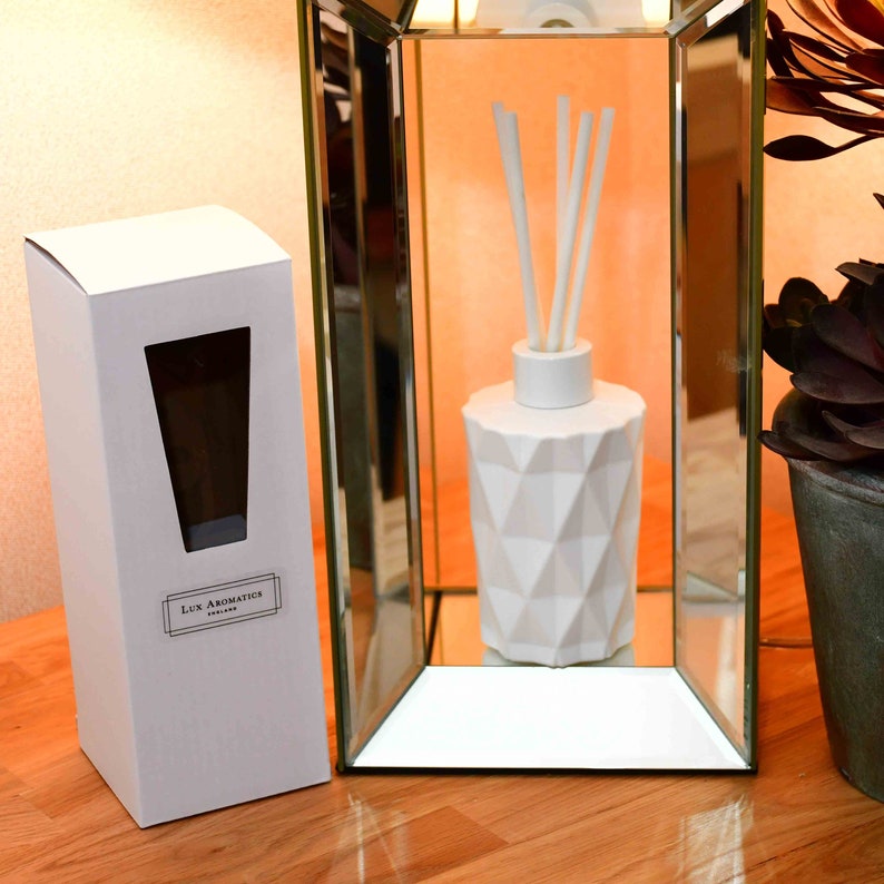 Puede incluir: Un difusor blanco con dise&ntilde;o geom&eacute;trico y varillas, exhibido en un marco espejado. A la izquierda, una caja blanca con ventana y el texto "Lux Aromatics England". Los objetos est&aacute;n sobre una superficie de madera.