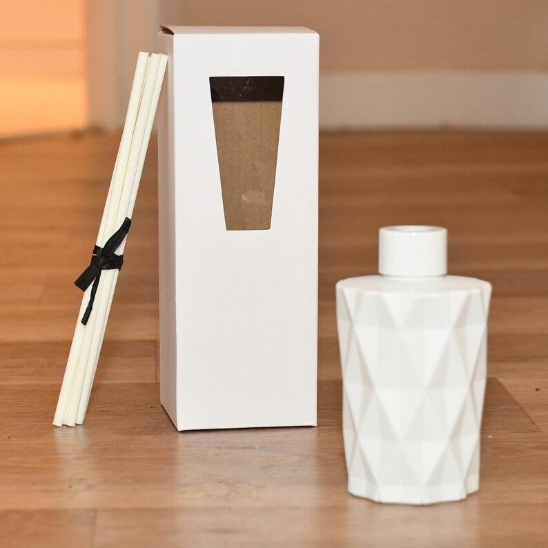 Puede incluir: Una botella difusora blanca con un patr&oacute;n geom&eacute;trico y una tapa a juego, una caja blanca con una ventana y un paquete de varillas difusoras atadas con una cinta negra. Sobre una superficie de madera.