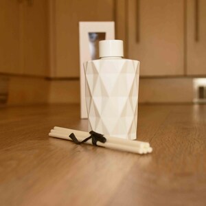 Puede incluir: Una botella difusora blanca con un patr&oacute;n geom&eacute;trico con una tapa a juego, un paquete de palitos difusores atados con una cinta negra y una caja blanca. La botella tiene un dise&ntilde;o de diamante. Los art&iacute;culos est&aacute;n sobre una superficie de madera.