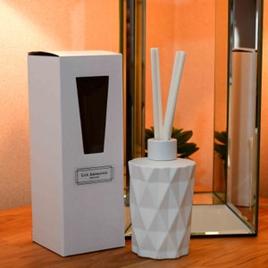 Puede incluir: Un difusor blanco con dise&ntilde;o geom&eacute;trico y varillas, junto a una caja blanca con ventana. El frasco tiene un dise&ntilde;o moderno. La caja tiene el texto "Lux Aromatics England". El fondo es un espejo y una pared de tonos c&aacute;lidos.