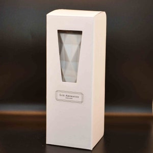 Puede incluir: Una caja rectangular blanca con una ventana que revela un recipiente de vidrio blanco con un patr&oacute;n geom&eacute;trico. La caja tiene el texto "Lux Aromatics England". El fondo es gris oscuro.
