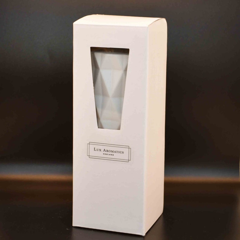 Puede incluir: Una caja rectangular blanca con una ventana que revela un recipiente de vidrio blanco con un patr&oacute;n geom&eacute;trico. La caja tiene el texto "Lux Aromatics England". El fondo es gris oscuro.