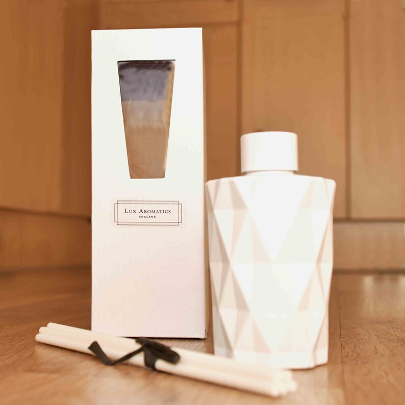 Puede incluir: Un difusor blanco con dise&ntilde;o geom&eacute;trico, una caja a juego y un manojo de varillas de ca&ntilde;a. La caja tiene una ventana que muestra un degradado de colores. El texto "Lux Aromatics England" est&aacute; impreso en la caja.