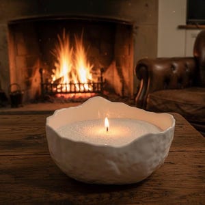 May include: A burning white candle in a textured, organic-shaped bowl on a wooden table. The candle's flame glows, with a fireplace and leather armchair in the background. The candle's wax has a granular texture.