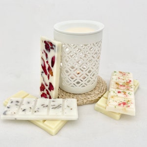 May include: A white ceramic wax melt burner with a decorative cut-out pattern, a small woven coaster, and several wax melt bars. The wax melts are white and decorated with dried flowers and herbs. The burner contains melted wax.