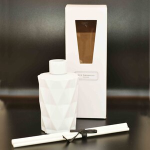Puede incluir: Un frasco difusor blanco con un patr&oacute;n geom&eacute;trico y una tapa a juego, presentado con una caja blanca y varillas. La caja presenta una ventana que revela un l&iacute;quido marr&oacute;n. El texto "Lux Aromatics ENGLAND" est&aacute; impreso en la caja. Una cinta negra asegura las varillas.