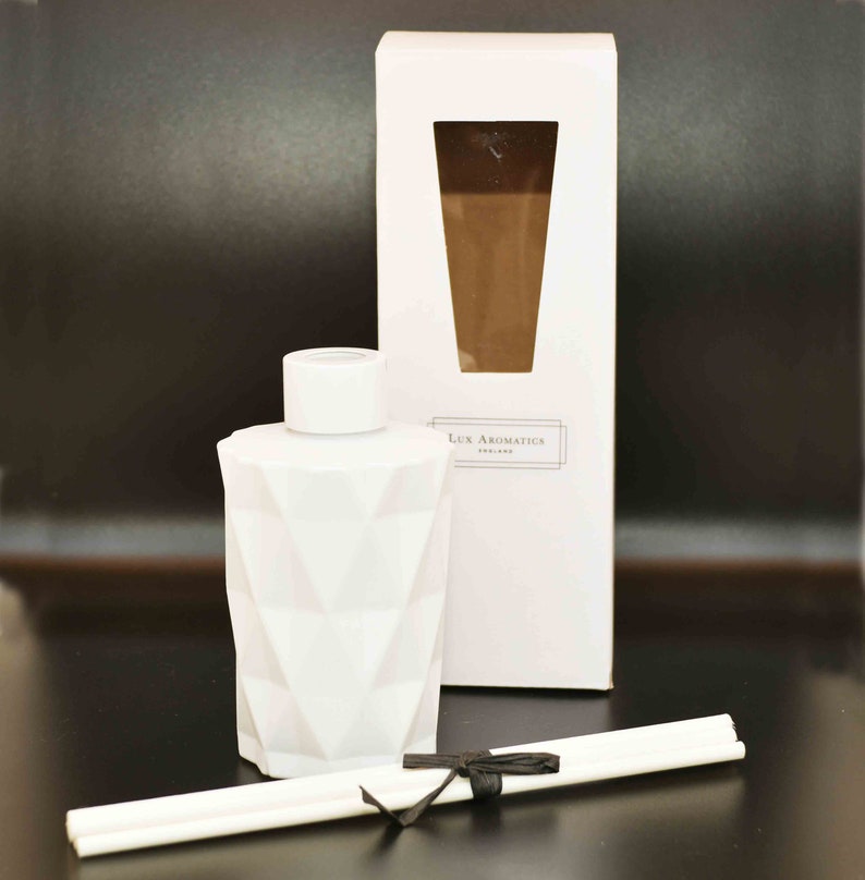 Puede incluir: Un frasco difusor blanco con un patr&oacute;n geom&eacute;trico y una tapa a juego, presentado con una caja blanca y varillas. La caja presenta una ventana que revela un l&iacute;quido marr&oacute;n. El texto "Lux Aromatics ENGLAND" est&aacute; impreso en la caja. Una cinta negra asegura las varillas.