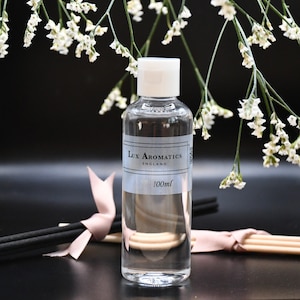 May include: A clear glass bottle of Lux Aromatics fragrance, labelled "ENGLAND" and "100ml", with a white cap. Black diffuser sticks, a pink ribbon, and white flowers are arranged around the bottle against a dark backdrop.