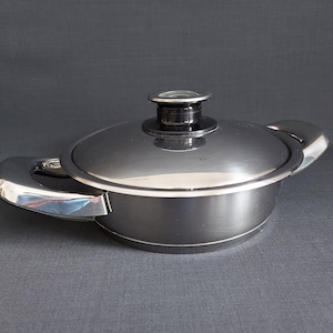 May include: A silver stainless steel saucepan with a lid and two handles. The lid has a black knob with a glass insert.