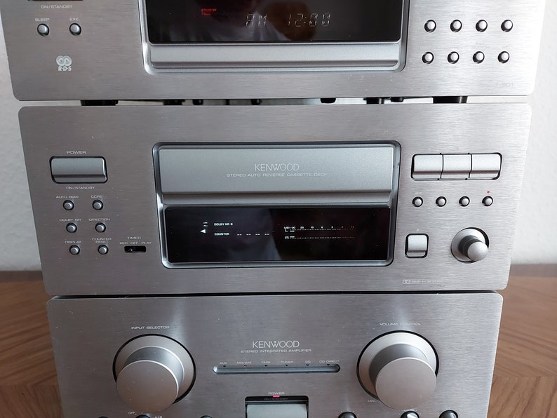Pu&ograve; includere: Un sistema stereo Kenwood argentato con un lettore CD, un registratore a cassette e un amplificatore. Il sistema ha un display digitale e diversi pulsanti per controllare le funzioni.