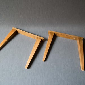 Op de afbeelding: Twee houten tafelpoten, elk met een rechthoekige bovenkant en schuine, taps toelopende poten. De poten zijn lichtbruin van kleur en lijken onafgewerkt. De poten staan tegen een grijze achtergrond.