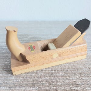May include: A wooden hand plane with a light brown finish. The plane features a curved handle, a blade, and a small sticker. This woodworking tool is designed for shaping and smoothing wood surfaces.