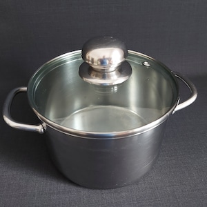 Könnte beinhalten: Ein Topf aus Edelstahl mit einem Glasdeckel und einem silbernen Knopf. Der Topf hat zwei Griffe.