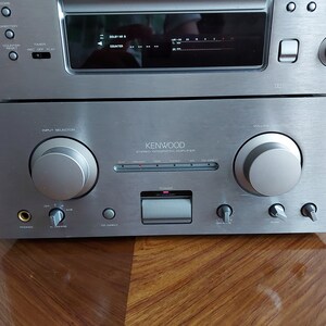 Pu&ograve; includere: Amplificatore integrato stereo Kenwood argentato con una piastra frontale argentata e manopole nere. L'amplificatore ha un pulsante di accensione, una manopola del volume, una manopola di selezione dell'ingresso e una variet&agrave; di altri controlli.