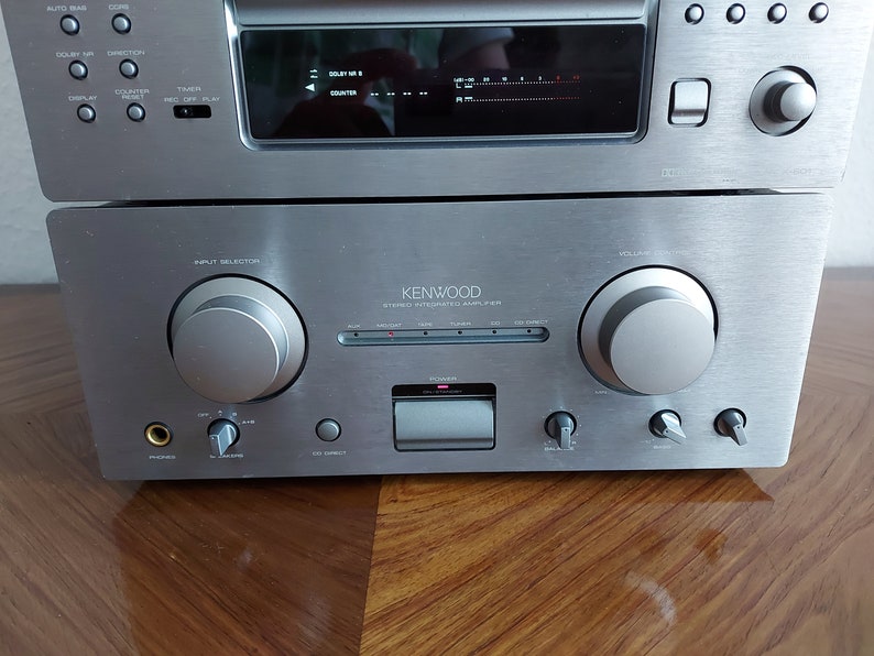 Pu&ograve; includere: Amplificatore integrato stereo Kenwood argentato con una piastra frontale argentata e manopole nere. L'amplificatore ha un pulsante di accensione, una manopola del volume, una manopola di selezione dell'ingresso e una variet&agrave; di altri controlli.