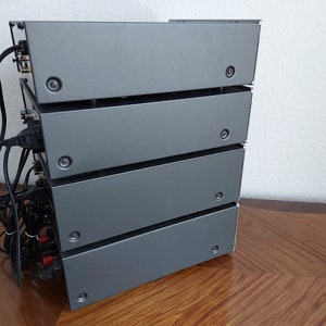 Pu&ograve; includere: Una pila di quattro componenti audio color argento con fili e connettori neri sul retro. I componenti sono impilati su una superficie di legno.