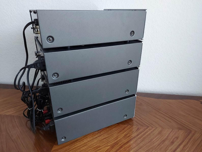 Pu&ograve; includere: Una pila di quattro componenti audio color argento con fili e connettori neri sul retro. I componenti sono impilati su una superficie di legno.