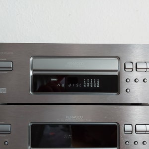 Pu&ograve; includere: Un lettore CD Kenwood argentato con un display digitale che mostra "no disc". Il lettore ha un vassoio di caricamento CD a slot e una variet&agrave; di pulsanti, tra cui accensione, apertura/standby e un pulsante di espulsione del disco. Il lettore &egrave; impilato sopra un ricevitore stereo Kenwood argentato con un display digitale e una variet&agrave; di pulsanti.