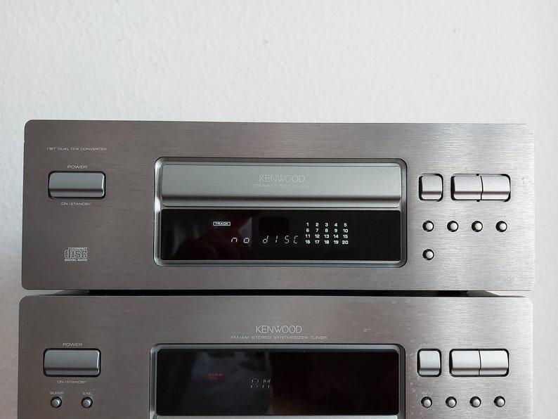 Pu&ograve; includere: Un lettore CD Kenwood argentato con un display digitale che mostra "no disc". Il lettore ha un vassoio di caricamento CD a slot e una variet&agrave; di pulsanti, tra cui accensione, apertura/standby e un pulsante di espulsione del disco. Il lettore &egrave; impilato sopra un ricevitore stereo Kenwood argentato con un display digitale e una variet&agrave; di pulsanti.